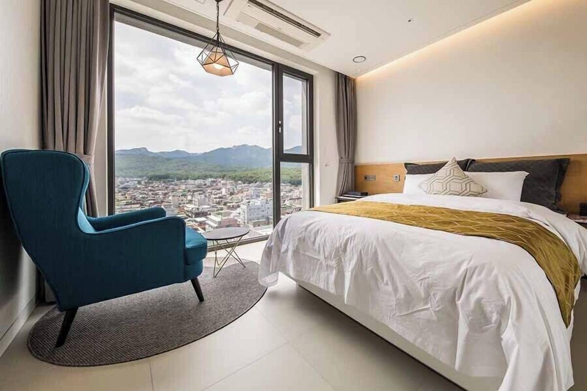 Ein Hotelzimmer mit einem großen Bett, einem blauen Sessel, Blick auf eine Stadtlandschaft durch ein großes Fenster und einem Deckenventilator.