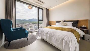 Une chambre d’hôtel avec un grand lit, un fauteuil bleu, une vue sur le paysage urbain par une grande fenêtre et un ventilateur de plafond.