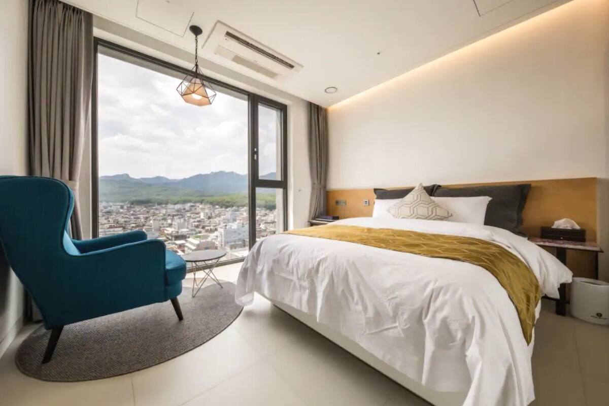 Ein Hotelzimmer mit einem großen Bett, einem blauen Sessel, einem kleinen Tisch und Blick auf eine Stadtlandschaft durch ein großes Fenster.
