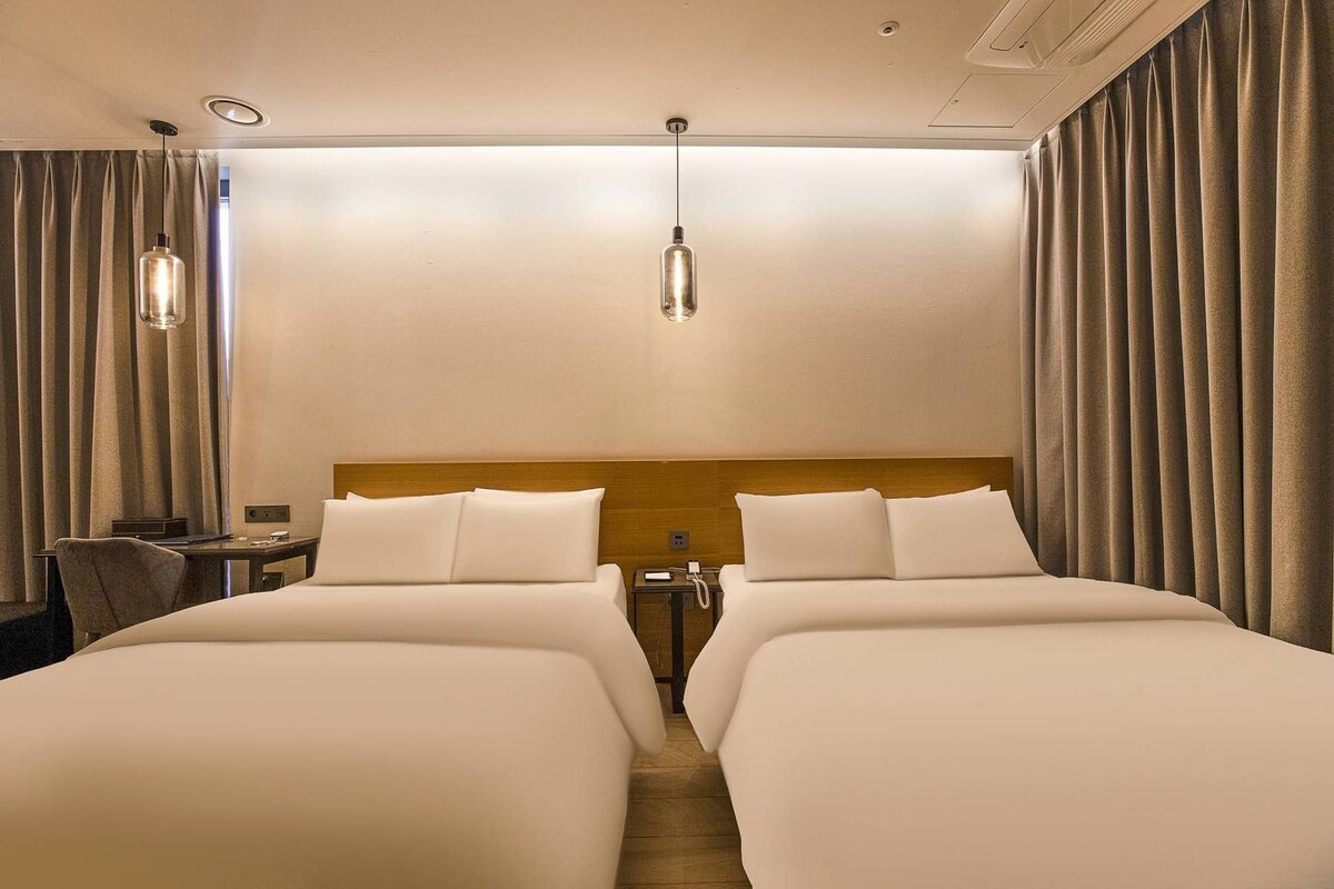 Une chambre d’hôtel avec deux lits, une tête de lit en bois, une table de chevet, une lampe et des rideaux.