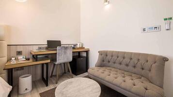 Um espaço compacto para viver, com sofá, mesinha redonda, escrivaninha com laptop, cadeira e lixeira.