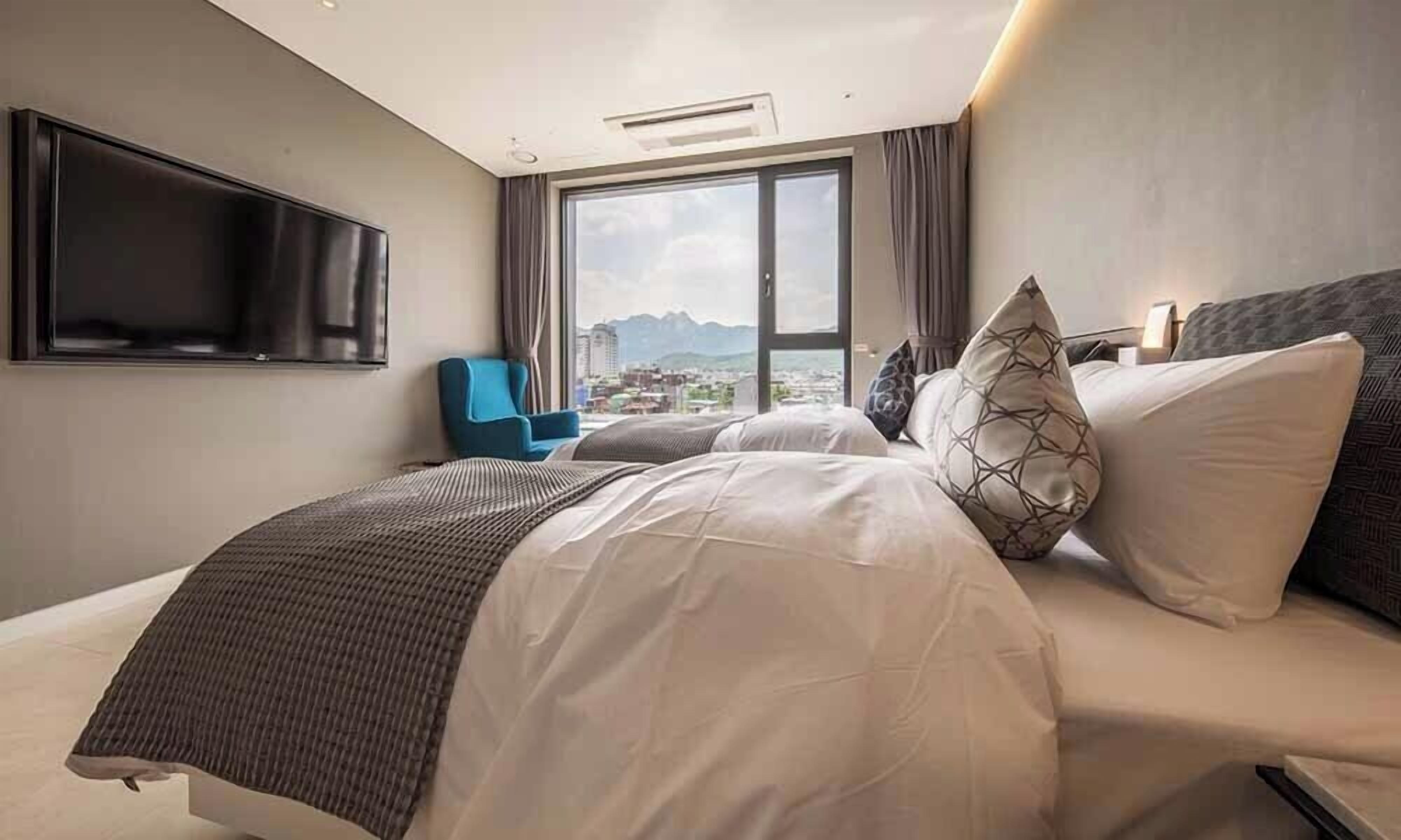 A hotel room with a large bed, a flat-screen TV mounted on the wall, a blue chair, and a view of a cityscape through a large window.