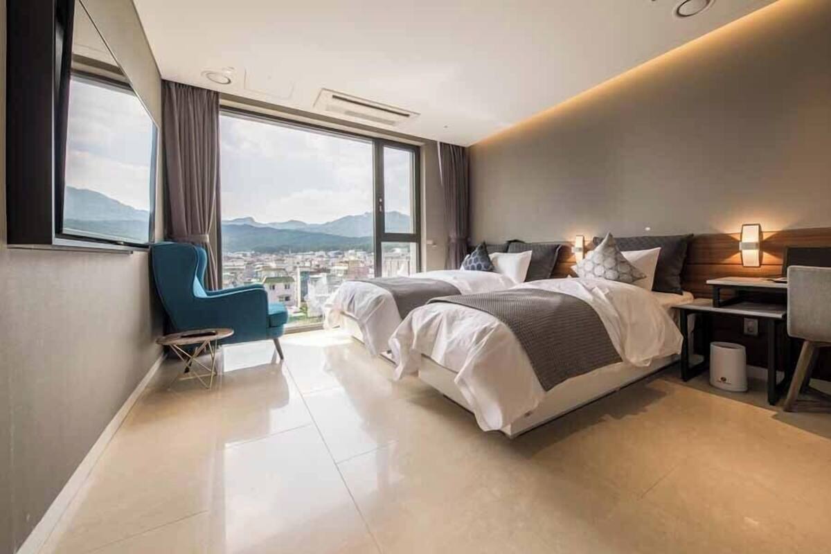 Ein modernes Hotelzimmer mit einem großen Bett, einem Schreibtisch, einem Sessel und Blick auf eine Stadtlandschaft durch große Fenster.