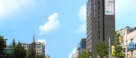Et hotel med flere etager og et tydeligt skilt, der siger 'Grand Open', beliggende på en travl gade med butikker og biler.