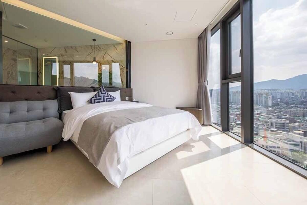 Ein modernes Schlafzimmer mit einem großen Bett, einer Sitzecke und Blick auf die Stadt durch ein großes Fenster.
