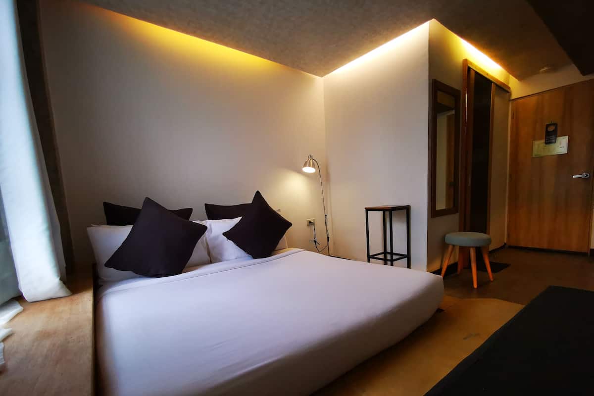 Un lit bien fait, avec une literie blanche et des oreillers noirs, une lampe de chevet, un petit tabouret et une porte en bois, dans une pièce aux murs blancs et au plafond en pente.