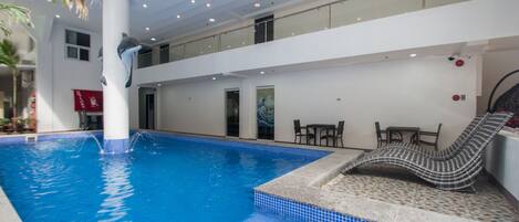 Una piscina interior con una escultura de delfín, un área de descanso con un tapete estampado y un techo alto con iluminación empotrada.