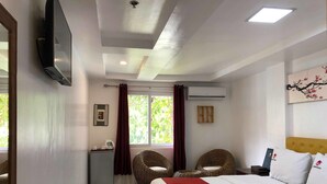 Una habitación de hotel con una cama, dos sillones de mimbre, una mesita, un televisor y un aire acondicionado.