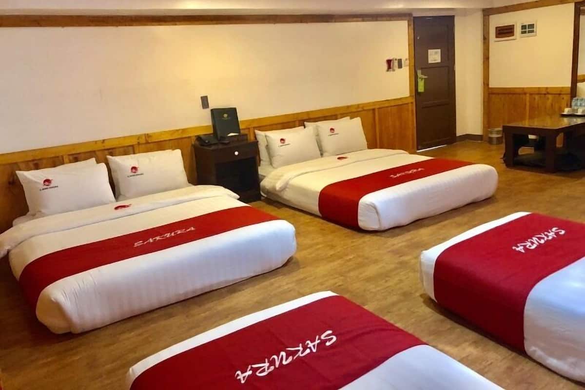 Habitación de hotel con dos camas, cada una con cubrecama rojo y blanco y almohadas a juego. Se observa un cabecero de madera, una mesita de noche con lámpara y una puerta a la derecha.