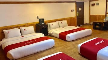 A hotel room with two beds, each with a red and white bedspread and matching pillows. There is a wooden headboard, a small bedside table with a lamp, and a door to the right.