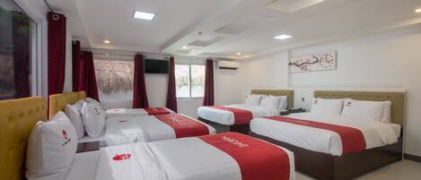 A hotel room with four beds, each with white bedding and red accents. There are large windows with red curtains, a ceiling with recessed lighting, and a wall-mounted artwork.