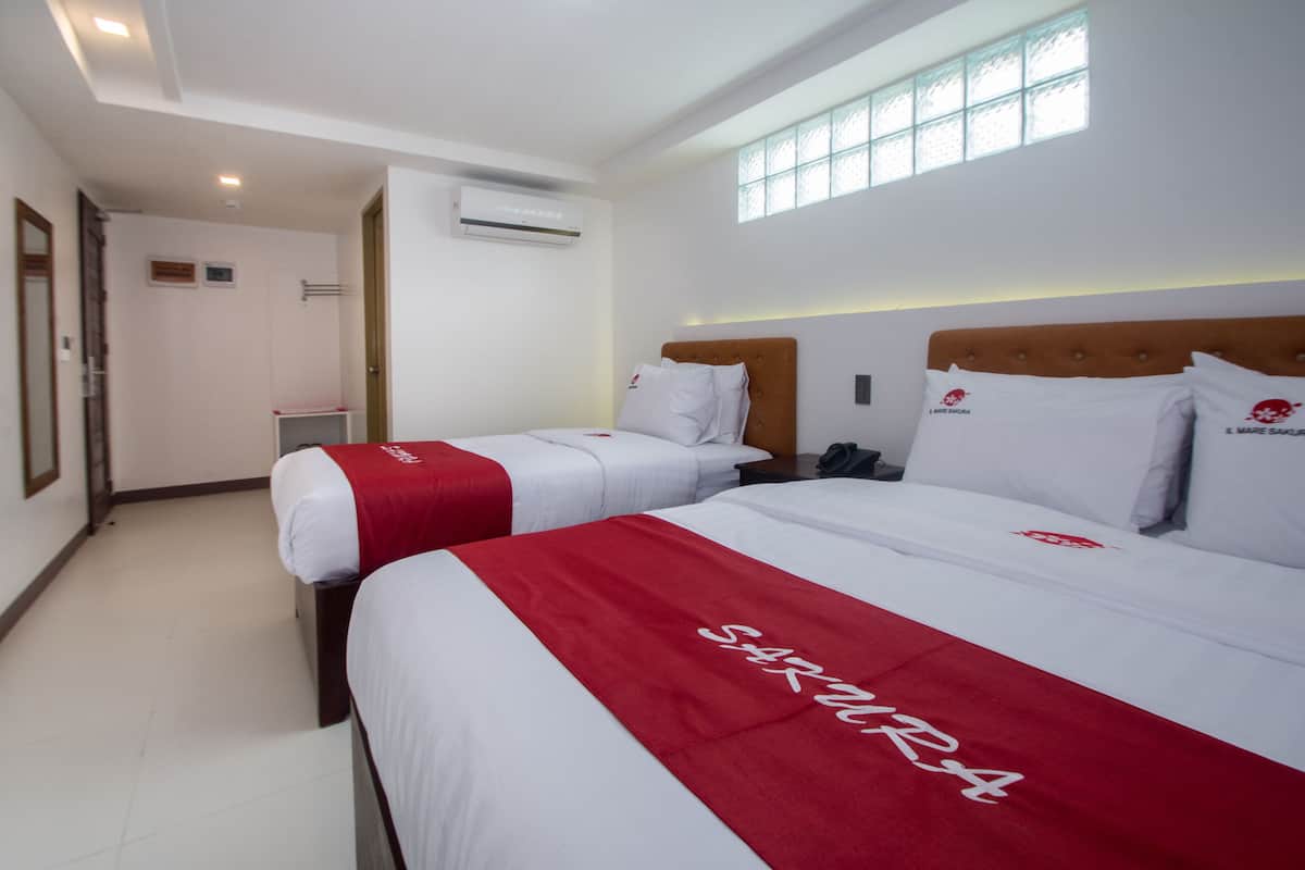 Habitación de hotel con dos camas, cada una con colcha roja y blanca, cabecero de madera y ventana que deja entrar luz natural.