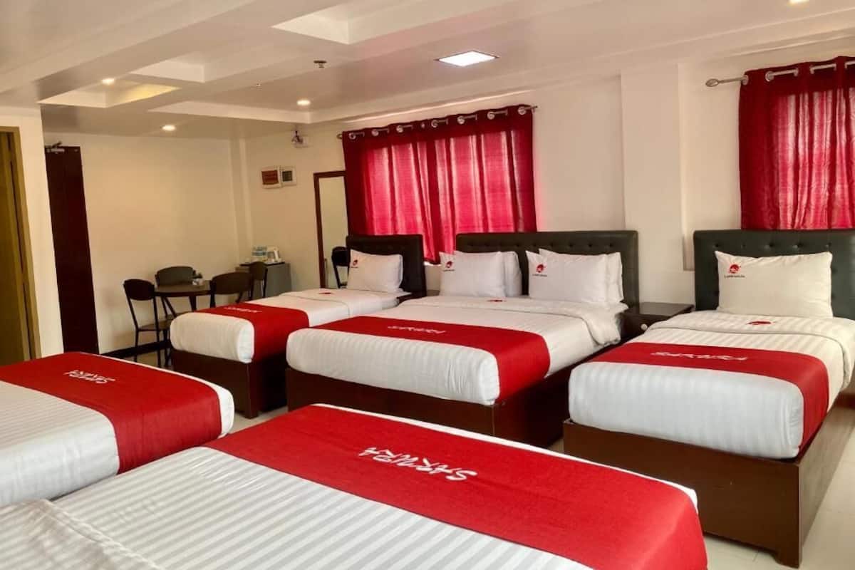 Habitación de hotel con tres camas, cada una con colcha y almohadas rojas y blancas. La habitación tiene un diseño moderno en el techo con luces empotradas y un ventanal con cortinas rojas.