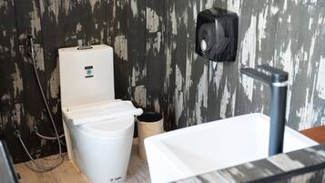 A bathroom with a white toilet, a trash can, a shower hose, and a soap dispenser on the wall.