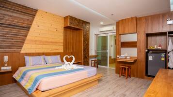 A modern hotel room with a wooden bed, a small kitchenette, a built-in wardrobe, and a sliding glass door leading to a balcony.