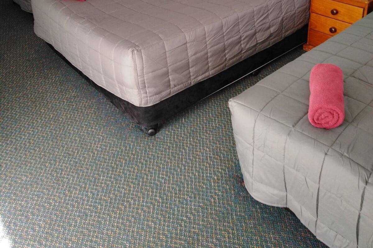 A hotel room with two beds, each with a grey mattress and red and white pillows. There is a wooden nightstand with drawers and a small table with a chair. The room has a blue wall and a carpeted floor.