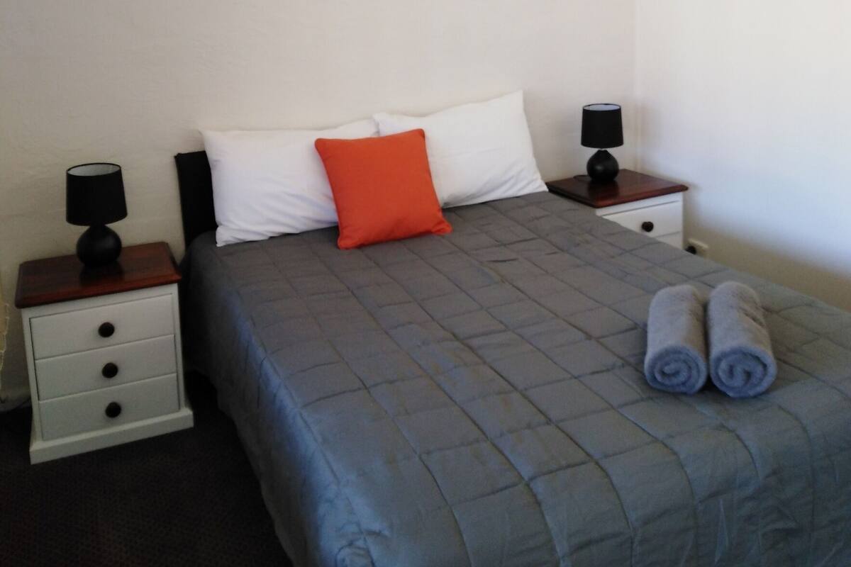A neatly made bed with a blue quilt, white and orange pillows, and two bedside lamps on white nightstands.
