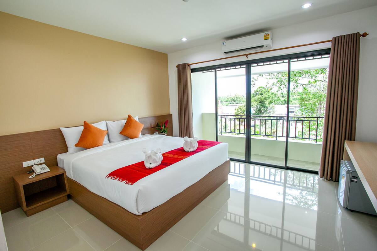 A hotel room with a large bed, a nightstand with a phone, a balcony with a view, and an air conditioning unit.