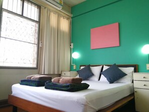 A hotel room with a bed, two bedside tables, a nightstand, a window with curtains, and a wall-mounted air conditioner. - 60 Bluehouse - Hostel (Chiang Mai)