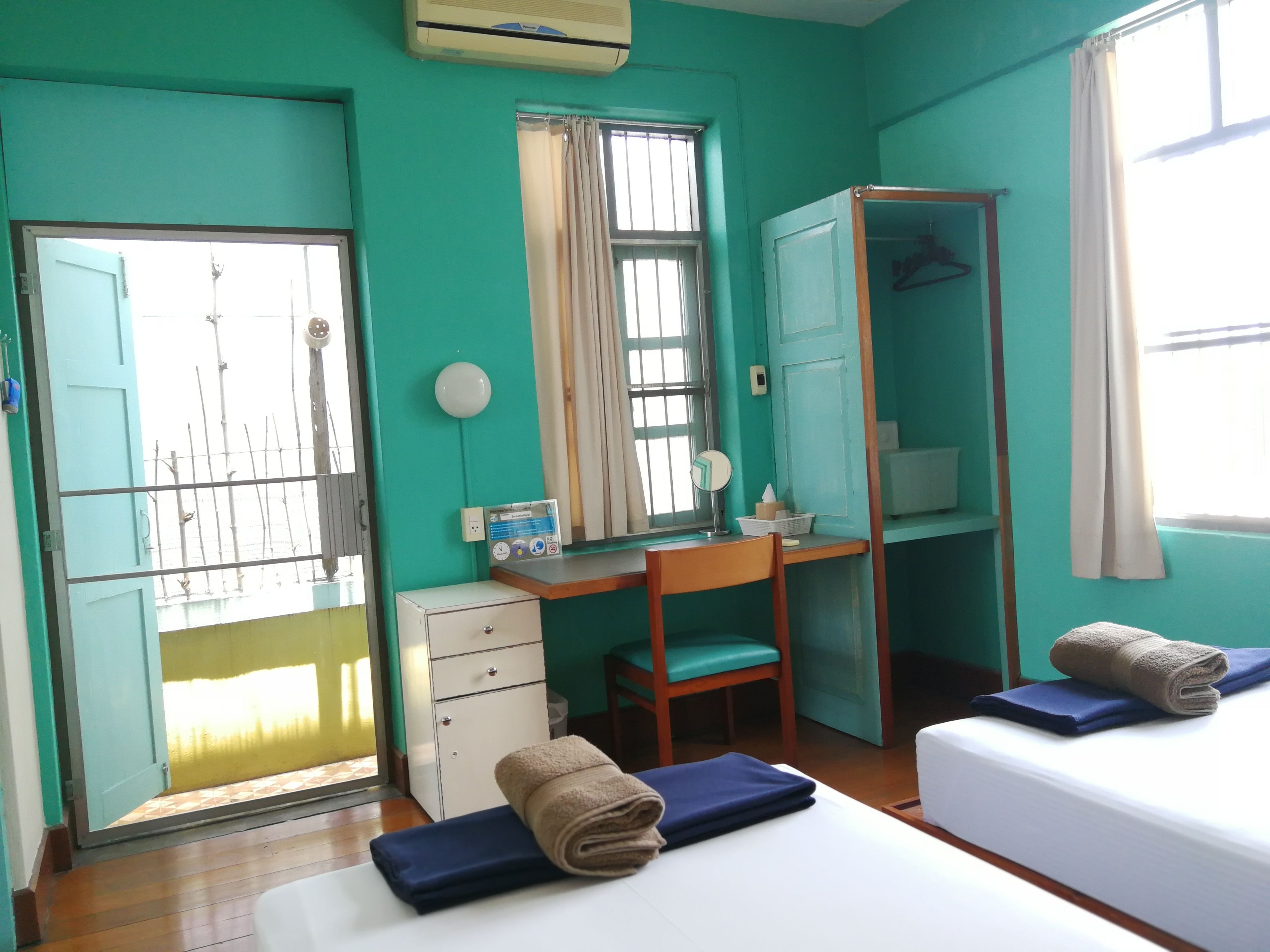 A room with teal walls, a desk with a chair, a wardrobe, a bed with two pillows, and a window with curtains.