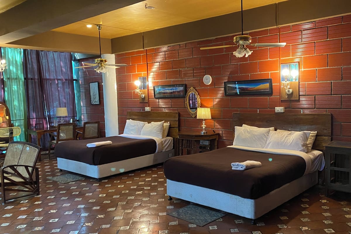 Habitación de hotel con dos camas, una pared de ladrillo, un ventilador de techo y un reloj.