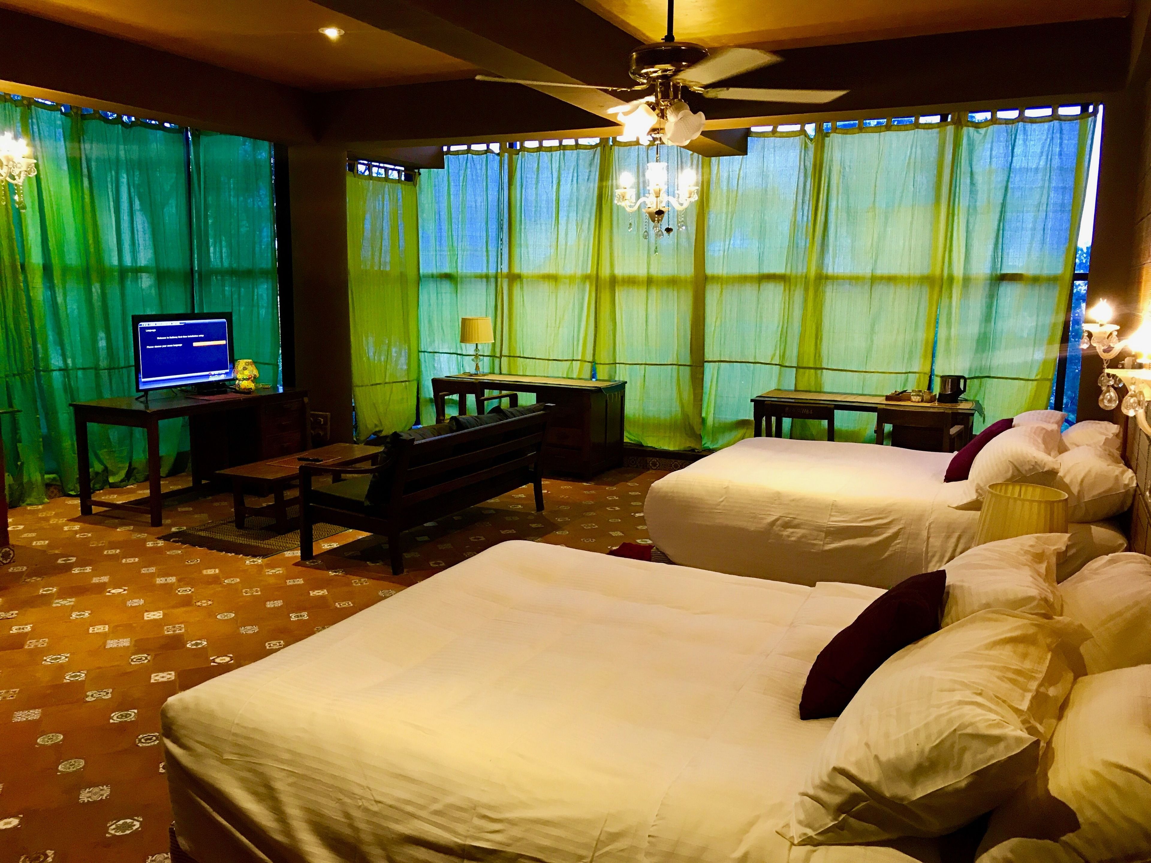 A hotel room with two beds, a desk, a chair, a television, and a ceiling fan.