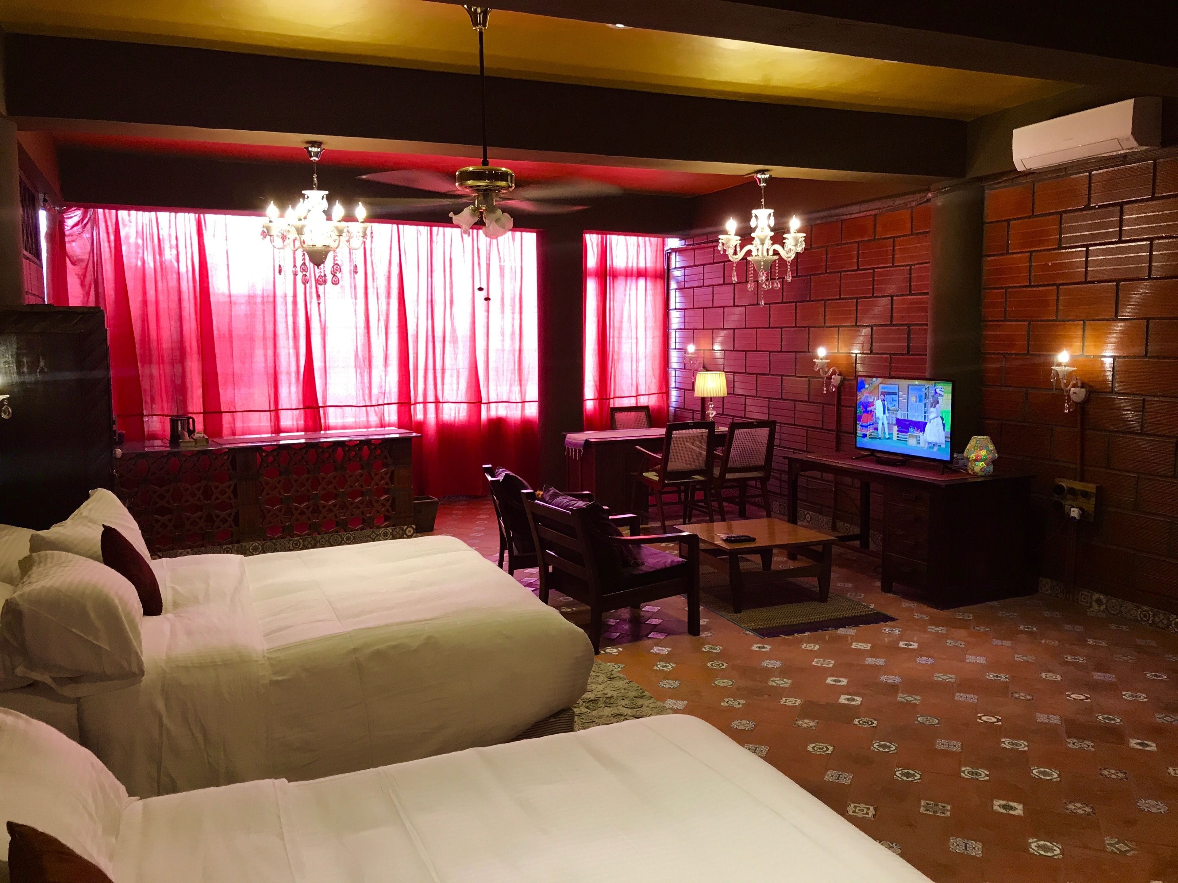A hotel room with two beds, a TV, a desk with a chair, and a brick wall.
