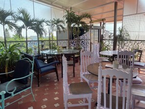 Un espacio al aire libre con un patio, mesas y sillas redondas, rodeado de plantas en macetas y una valla decorativa.