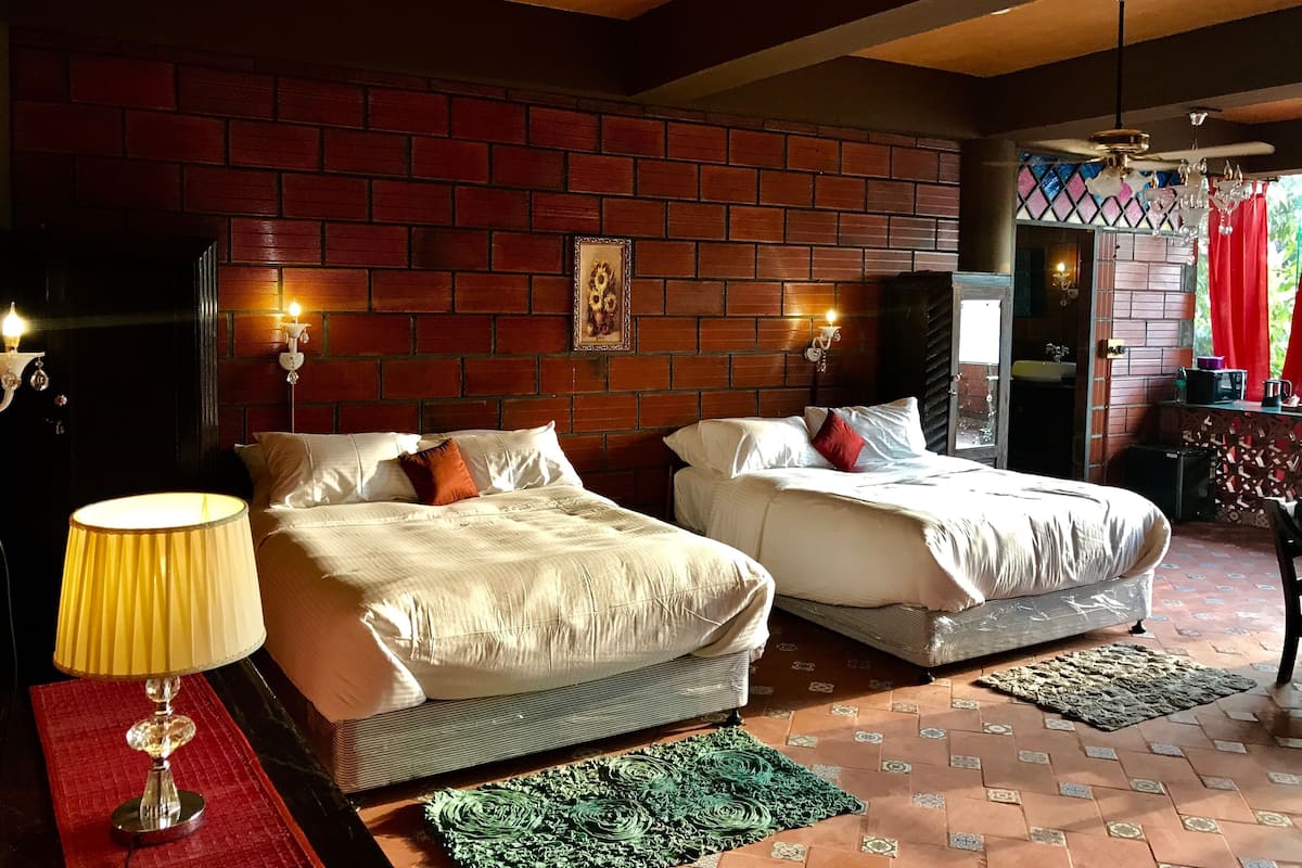 Una habitación con dos camas, una pared de ladrillo, una araña y una ventana con cortinas rojas.