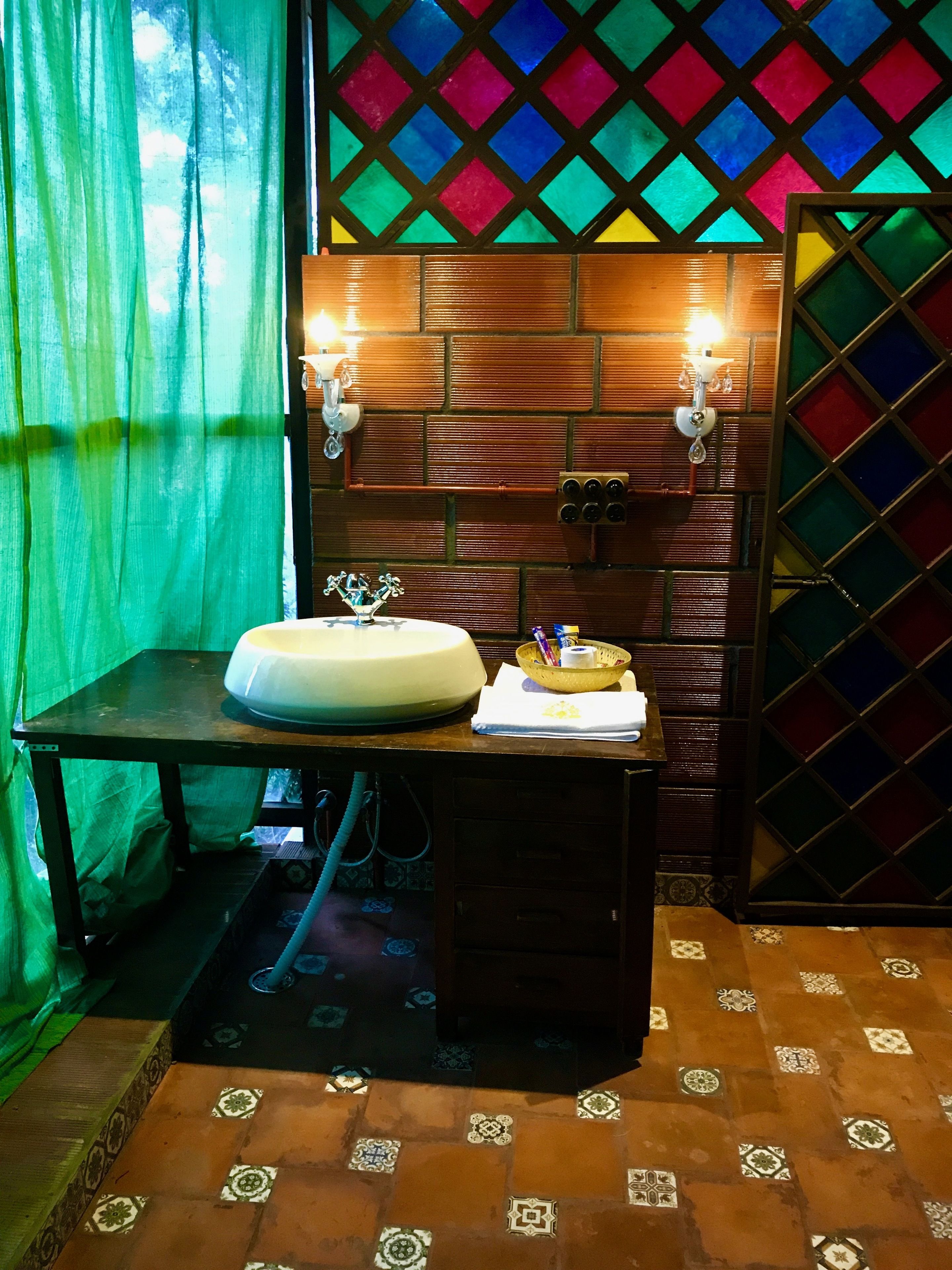 A bathroom with a wooden vanity, a sink, and a mirror. There are two wall-mounted lights and a colorful stained glass window.