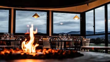 Un restaurante con un elemento decorativo de fuego, mesas dispuestas para comer y amplias ventanas que ofrecen vistas al exterior.