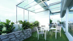 A rooftop terrace with a glass enclosure, green artificial grass, a table with chairs, and a variety of plants. - Marino Royal Hotel (Dhaka)
