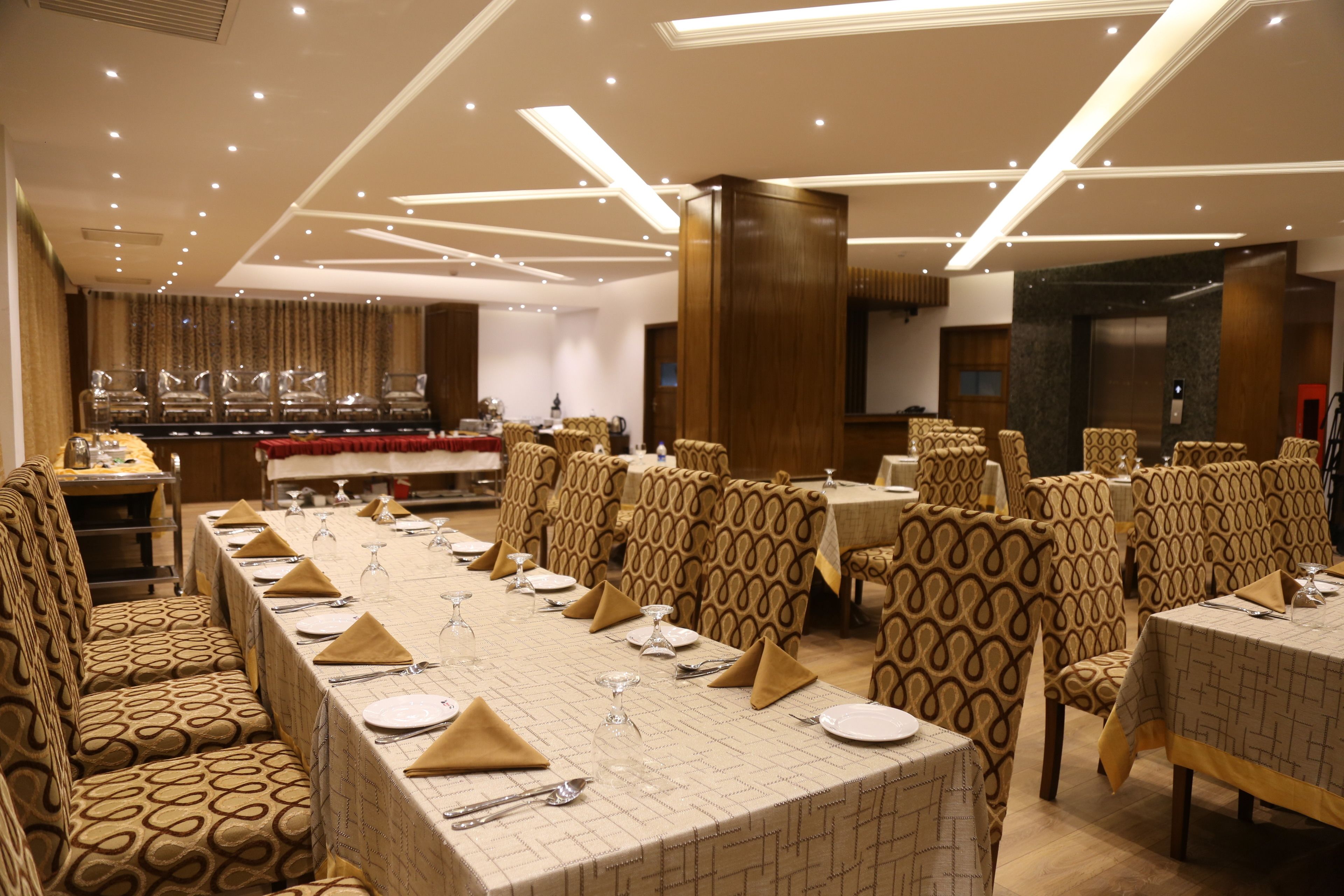 Une salle de banquet avec de longues tables dressées pour le repas, des chaises à motifs et un design de plafond moderne.