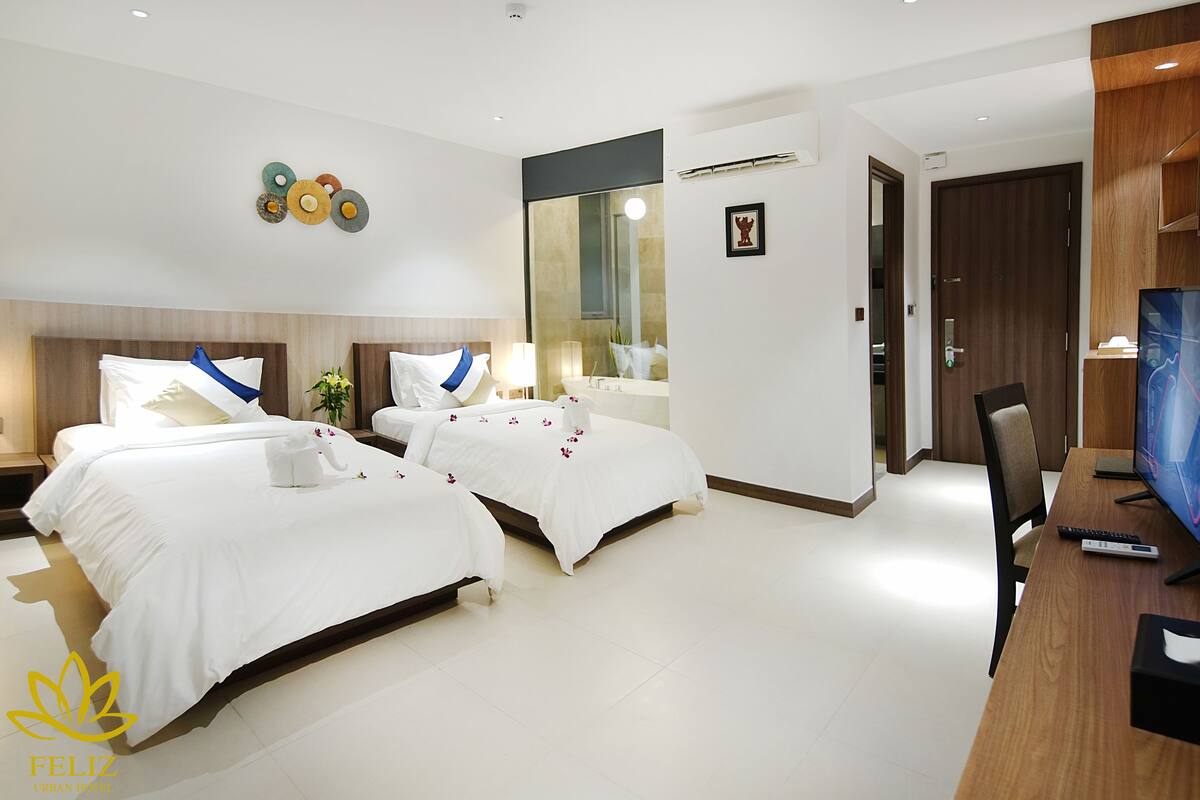A hotel room with two beds, a desk with a computer, a TV, and a bathroom with a mirror and sink.