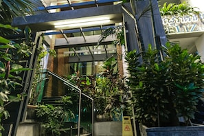 A modern building entrance with glass railings, a wet floor sign, and lush indoor plants. - Feliz Urban Hotel (Phnom Penh)