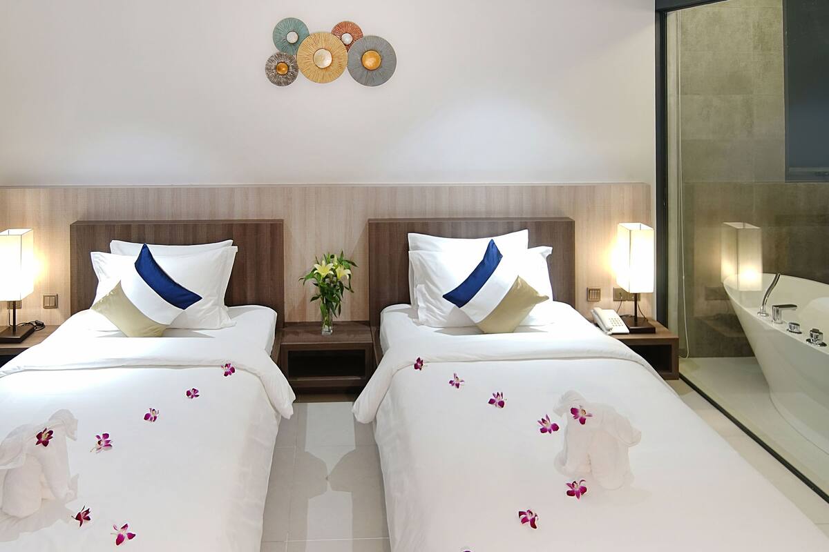 A hotel room with two beds, each with a nightstand, a lamp, and a vase with flowers. There is a large mirror and a bathtub visible.