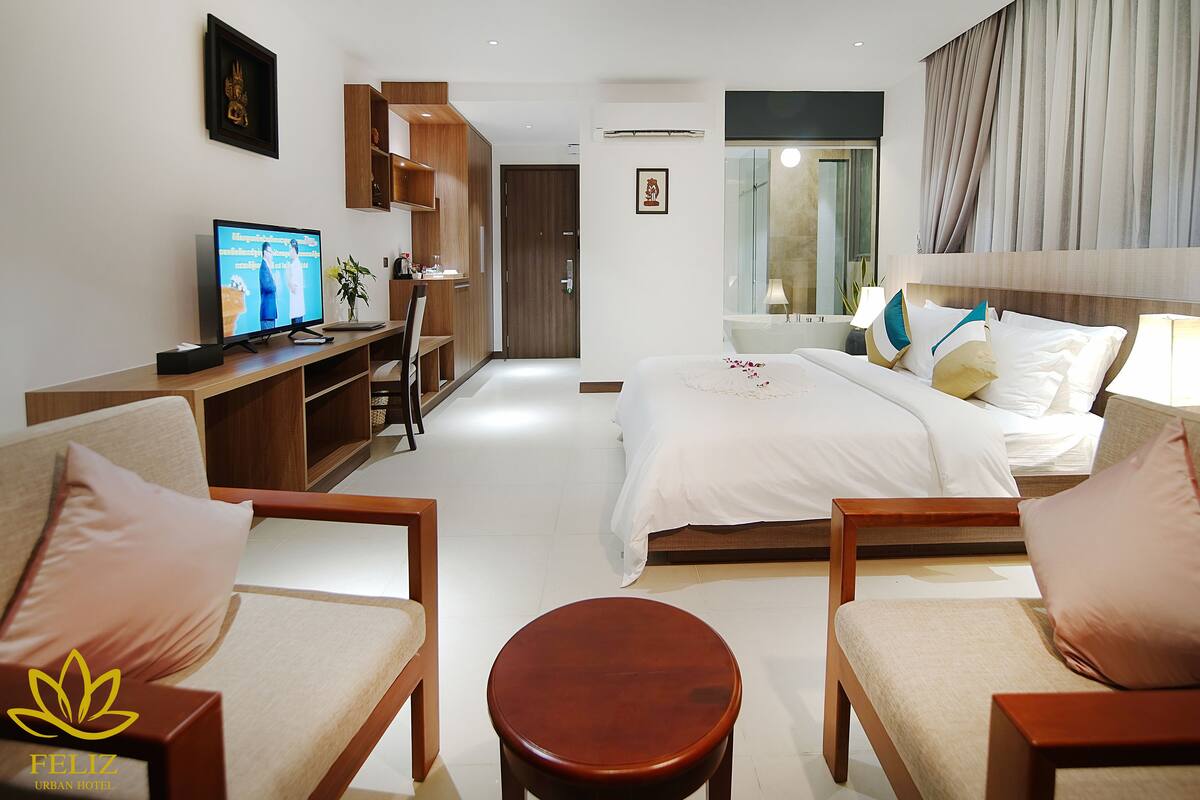 A modern hotel room with a bed, two armchairs, a wooden side table, a television, and a desk with a chair.