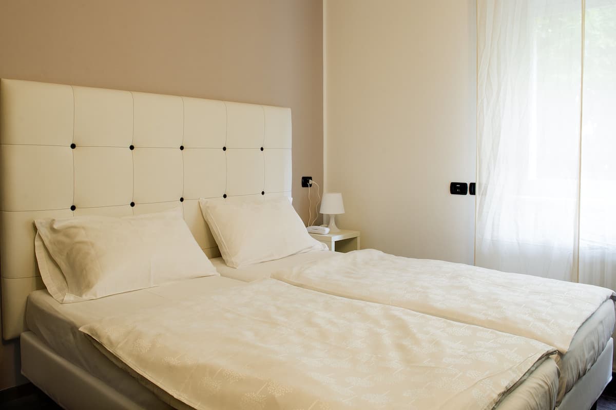 Una cama doble con cabecera capitonada, una mesita de noche con lámpara y una puerta con cortina.