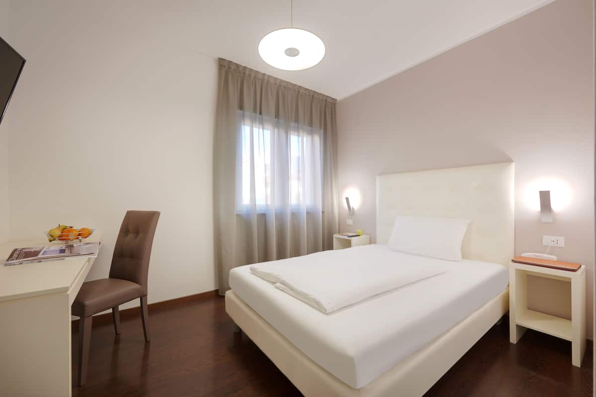 Una habitación de hotel con cama, escritorio, silla, mesita de noche y ventana con cortinas.