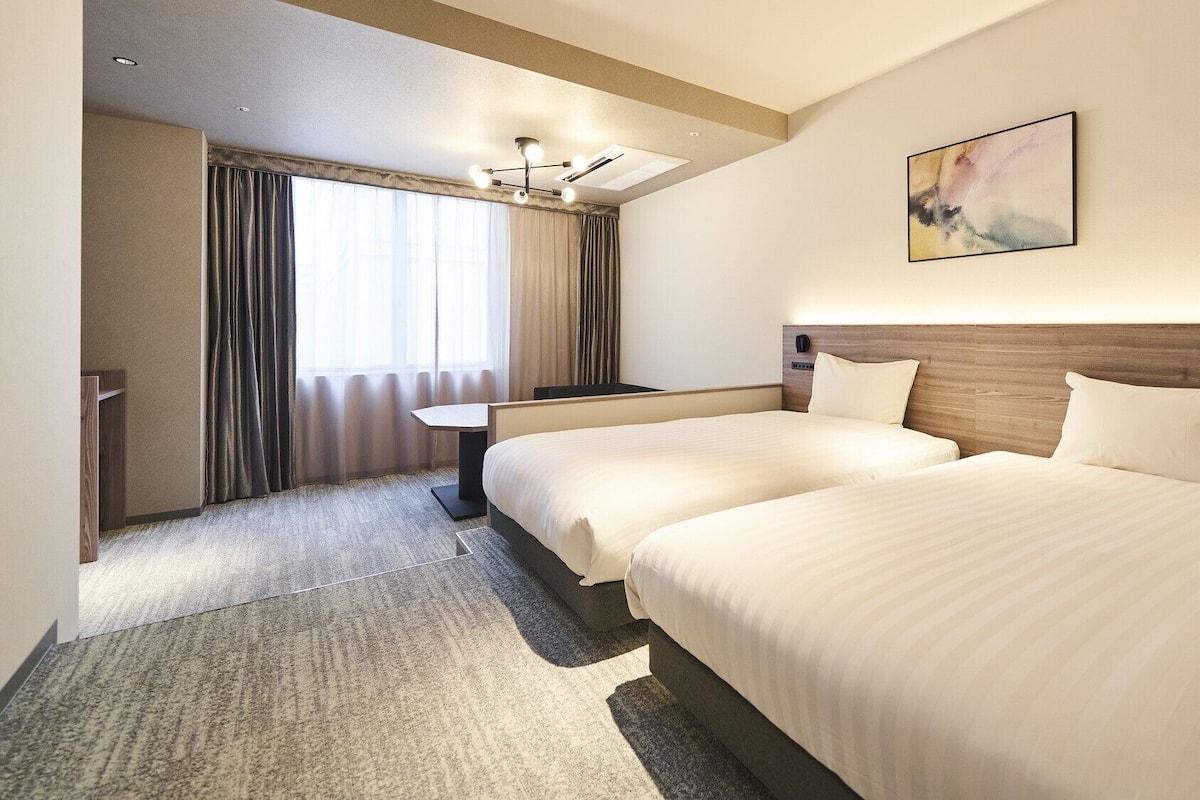 A hotel room with two beds, a desk, a chair, a painting on the wall, and a window with curtains.