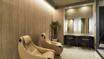 A spa room with two massage chairs, a potted plant, and a mirror above a table with two chairs.