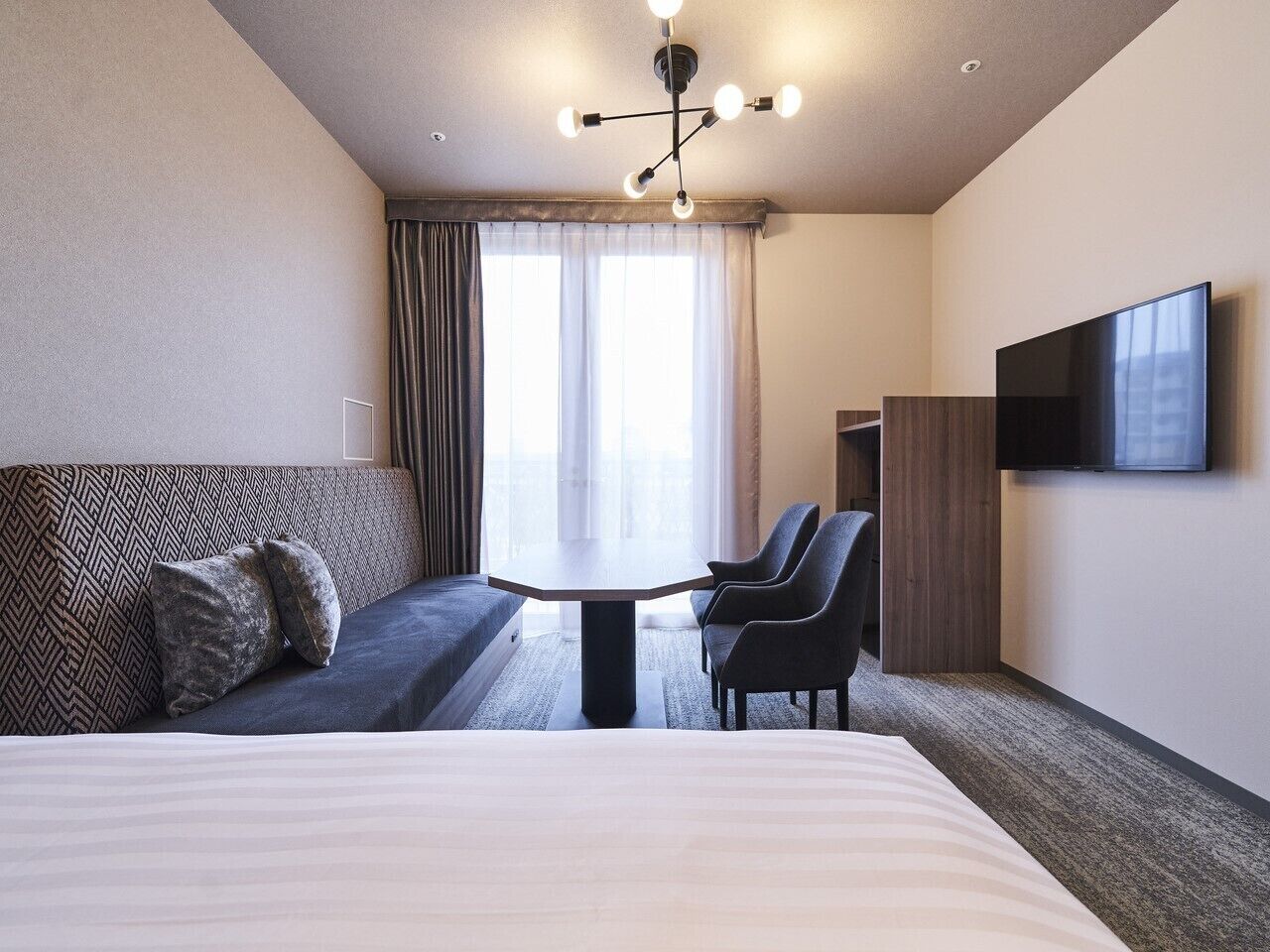 A hotel room with a large bed, a sofa, a dining table with chairs, a TV, and a window with curtains.