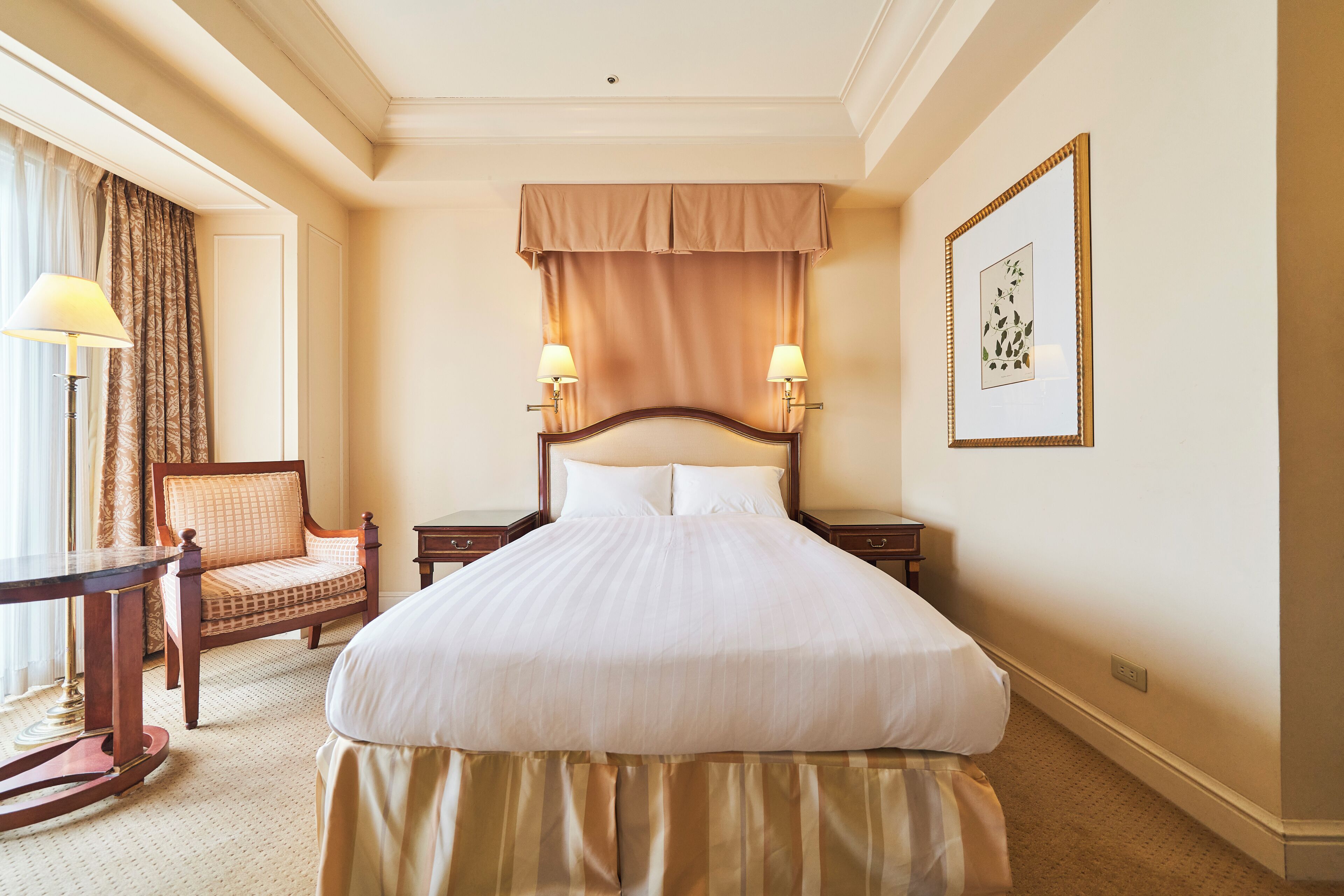 Habitación de hotel con una cama grande, dos lámparas de noche, una silla, una mesa y un cuadro en la pared.