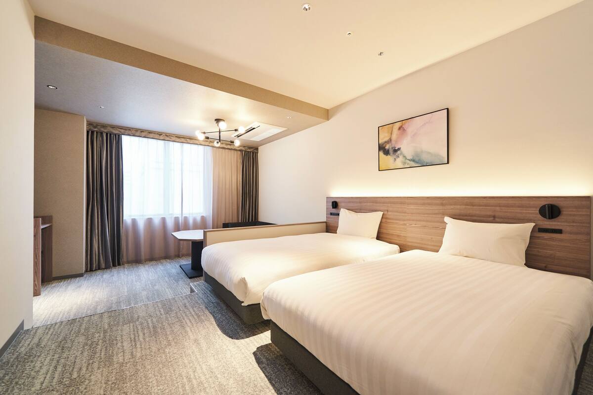 A hotel room with two beds, a desk, a chair, a large window with curtains, and a ceiling light fixture.