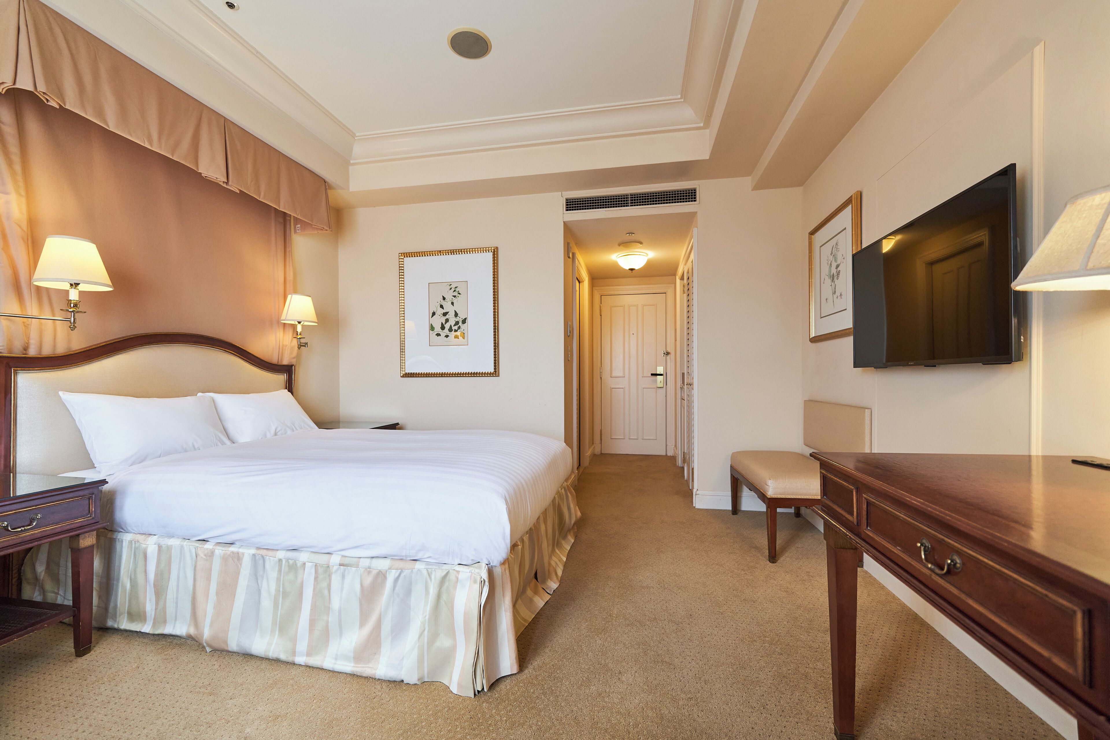 A hotel room with a large bed, bedside lamps, a desk, a television, and a framed picture on the wall.