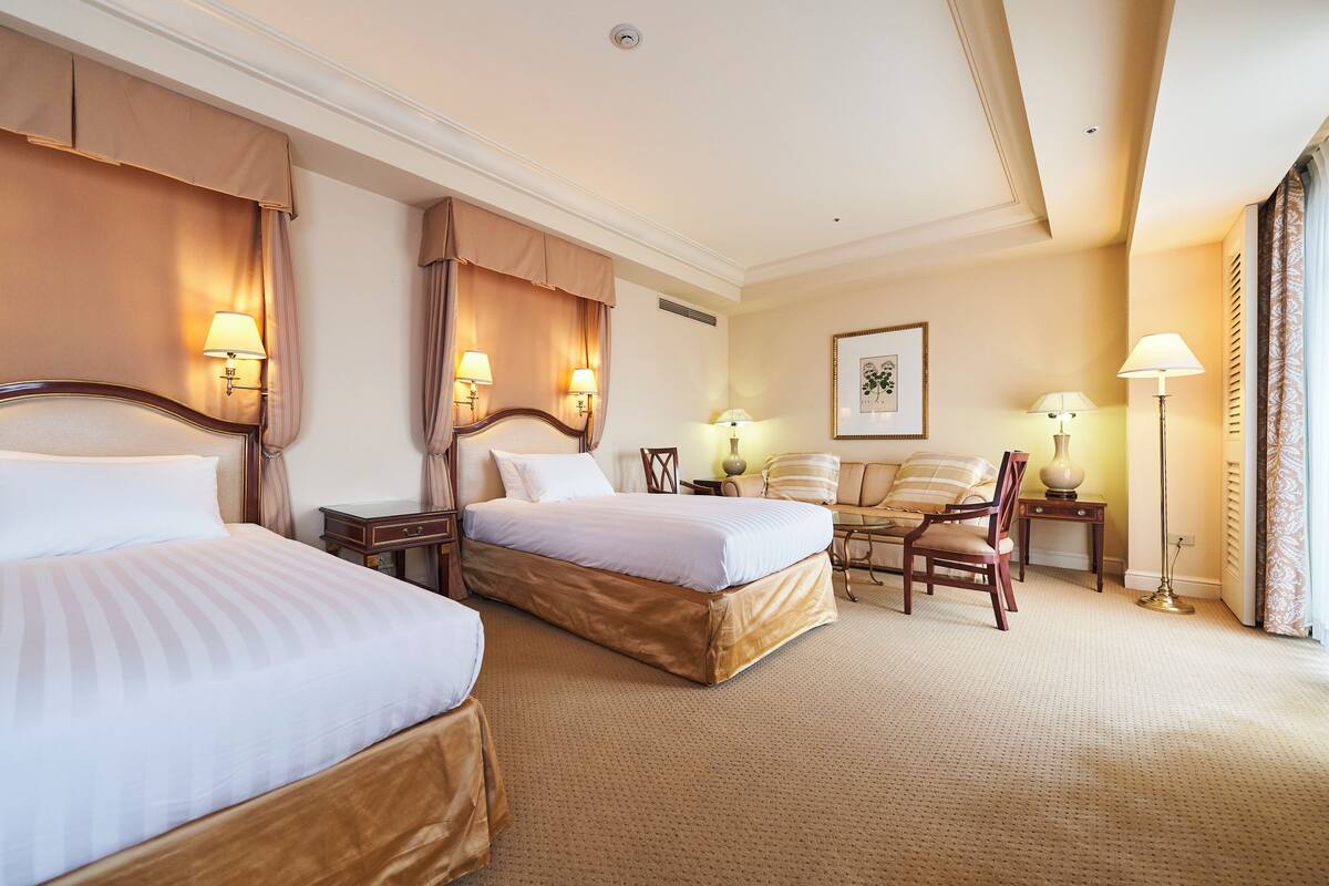 A hotel room with two beds, a desk, a chair, and a lamp.