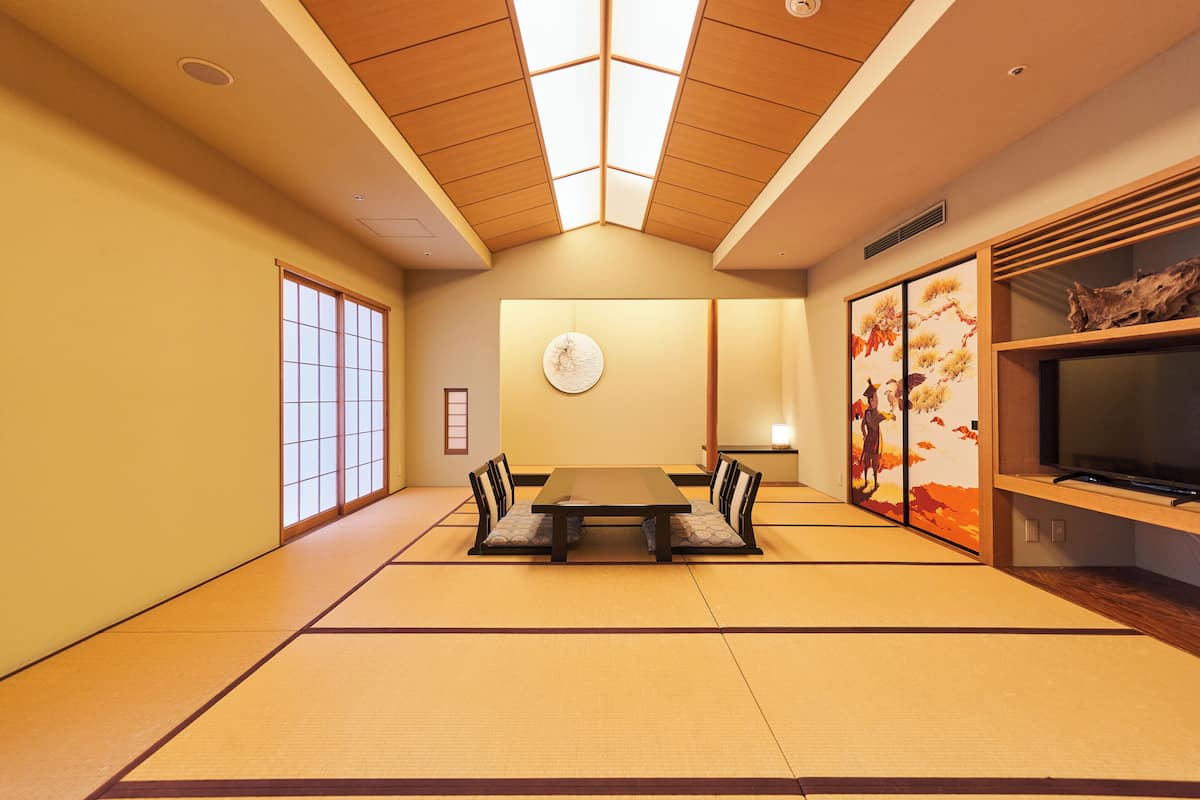 A traditional Japanese room with tatami flooring, a low table with cushions, a television, and artwork on the walls.