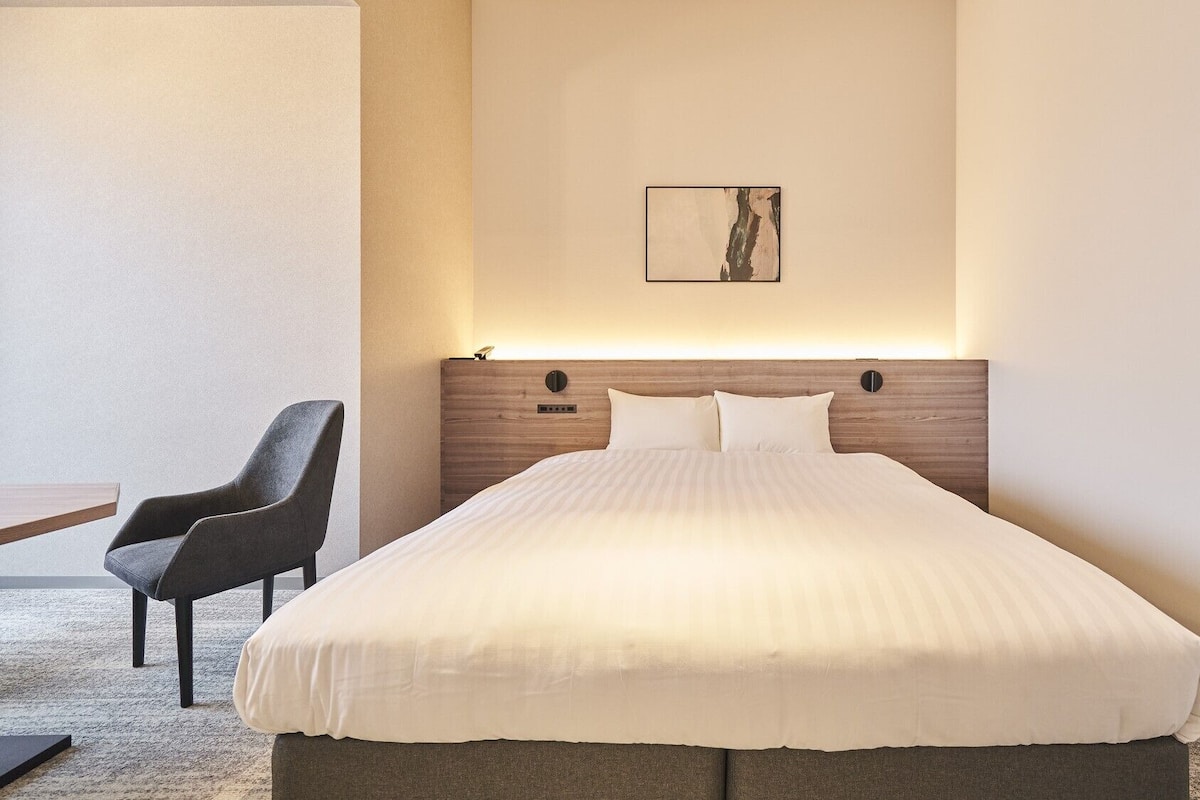 A hotel room with a bed, a chair, a bedside table, and a wall-mounted artwork.