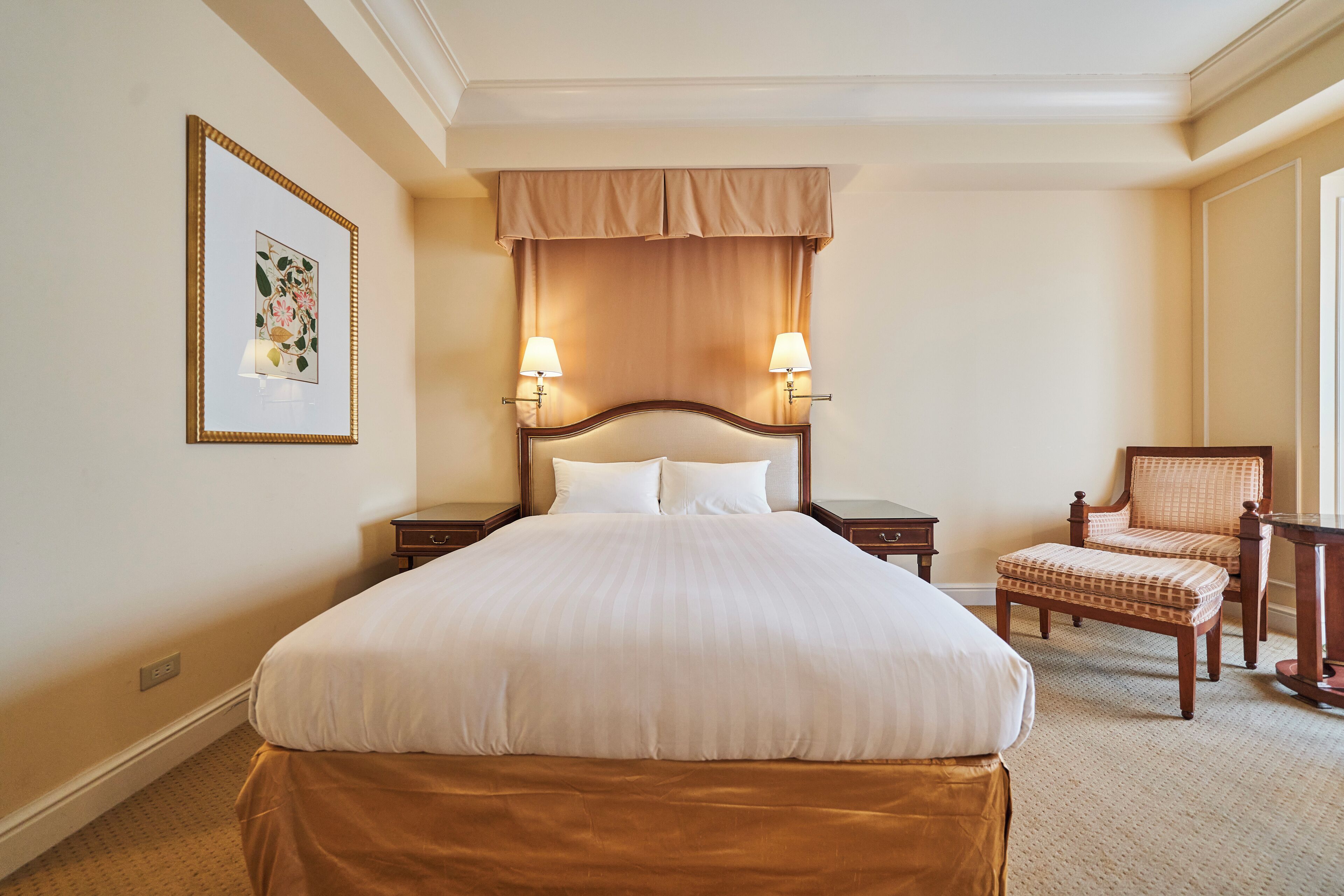 A hotel room with a large bed, two bedside lamps, a chair, and a framed picture on the wall.