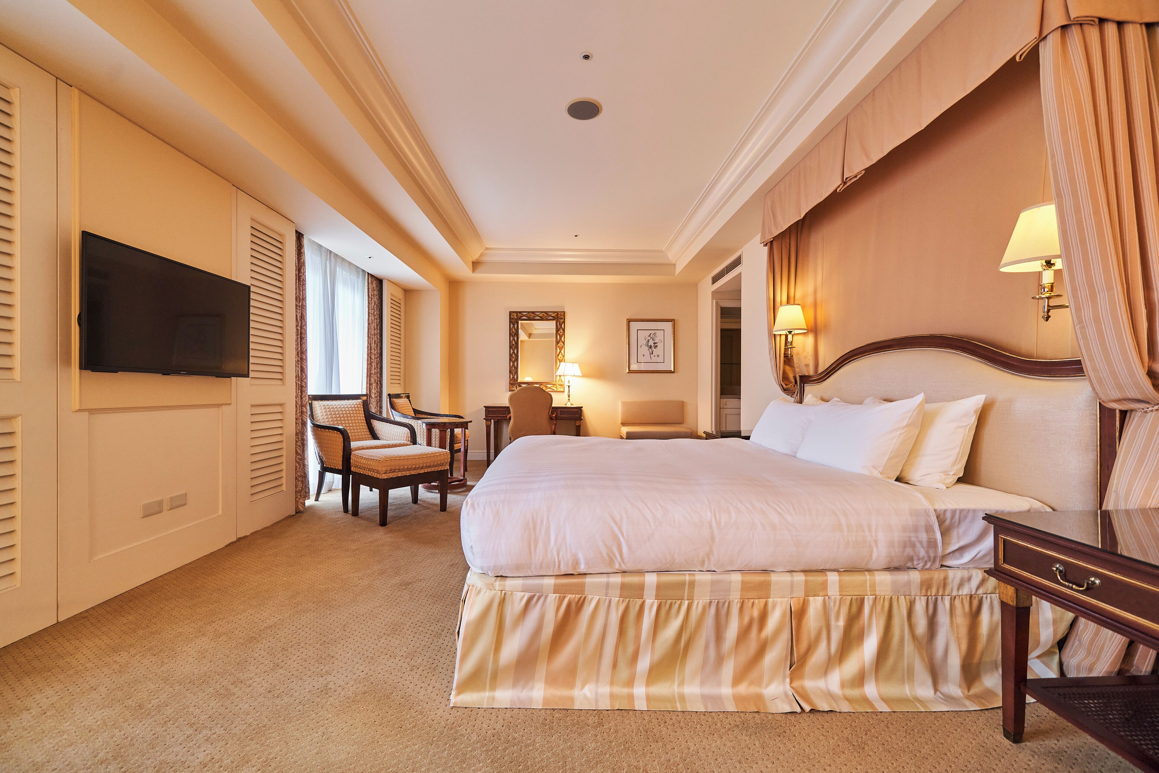 A hotel room with a large bed, a television mounted on the wall, a desk with a lamp, and a chair.
